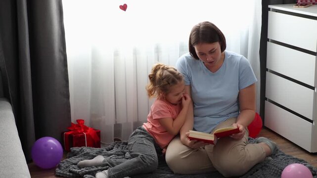 Caucasian mother reading a storybook with her young daughter while relaxing on the floor in a cozy living room. Early childhood education and family time concept. Development through reading and commu