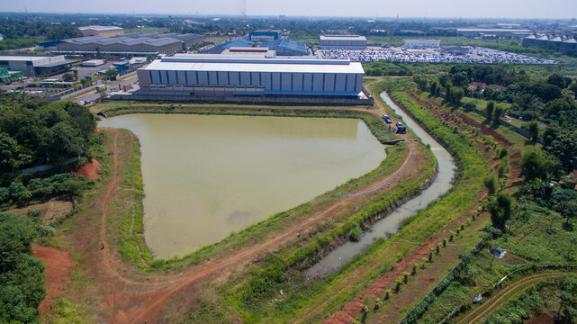 Aerial view of a large industrial warehouse facility situated next to a retention pond and surrounded by lush green fields and vegetation in Cikarang Barat, West Java, Indonesia.
