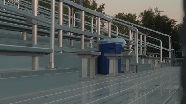 Slow right to left pan across empty aluminum stadium bleachers with a blue recycling bin.