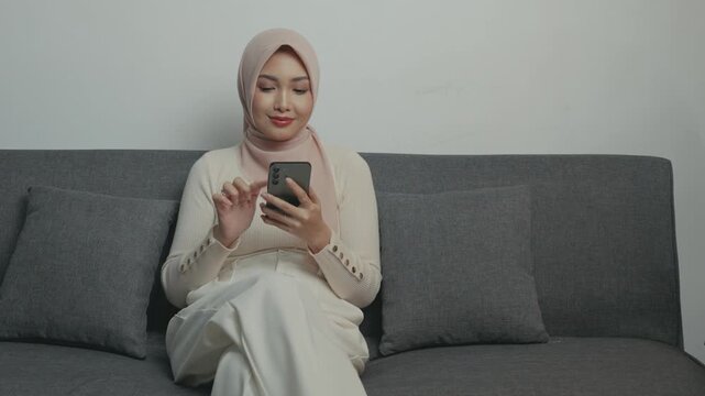Pretty young Asian Muslim woman in hijab using smartphone while sitting down on a sofa.
