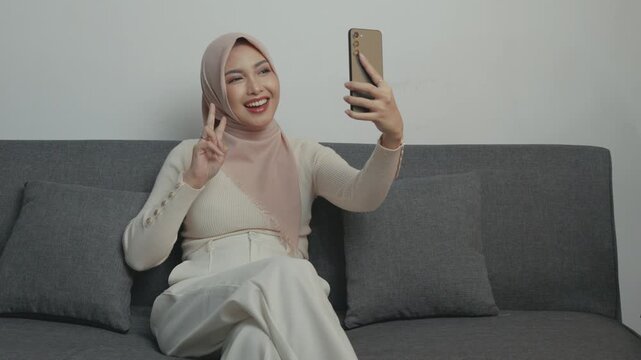 Smiling young Muslim Asian woman in a hijab is using smartphone while taking pictures of herself.