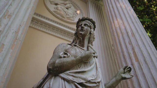 Medium shot of a weathered marble statue of a classical female figure adorned with a flower crown and holding a pan flute, against a decorative relief wall of a historic structure