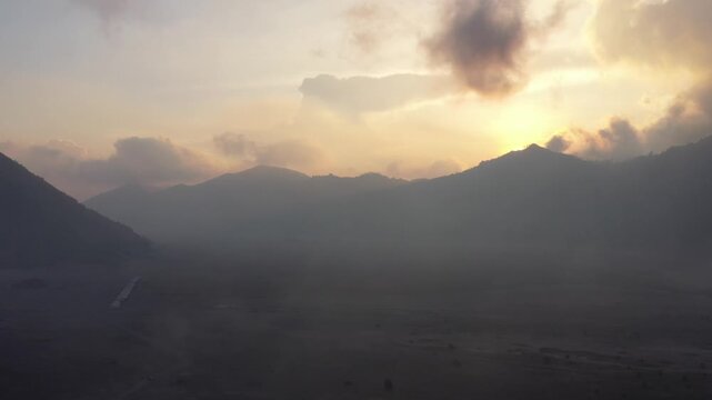 Aerial drone pans left to reveal Mount Batok and smoking Mount Bromo at sunrise, low clouds over the caldera, East Java, Indonesia. Cinematic volcanic landscape b-roll.