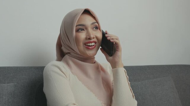 Joyful young Asian woman wearing hijab smiling and having a call with someone while sitting on a sofa.