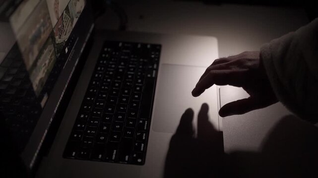 Close-up of hand operating laptop trackpad in low-light home office. Concept of overtime work, focus and modern remote digital workflow.