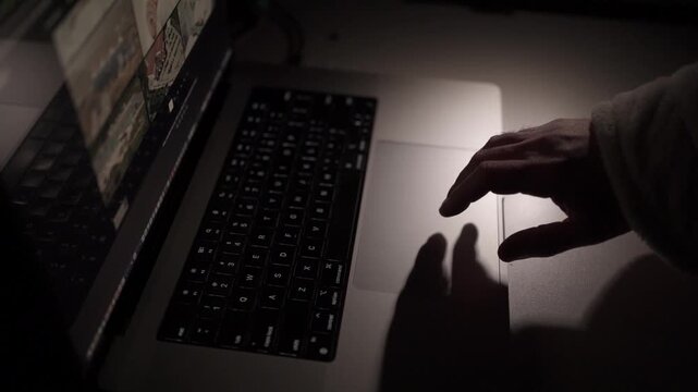 Close-up of hand operating laptop trackpad in low-light home office. Concept of overtime work, focus and modern remote digital workflow.