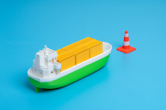 Green cargo ship stopped by an orange traffic cone. Shipping delays, supply chain disruptions, port congestion, maritime trade blockades, and logistics warnings concept..