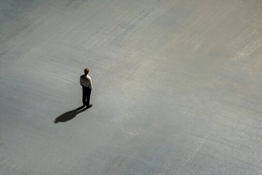 A solitary miniature businessman stands on a vast, textured gray surface, casting a long shadow. Social isolation, uncertainty, daunting business challenges, loneliness concept.