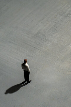 A solitary miniature businessman stands on a vast, textured gray surface, casting a long shadow. Social isolation, uncertainty, daunting business challenges, loneliness concept.