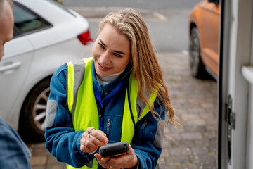 Delivery worker interacts with customer in residential area while checking details on handheld device © Iryna