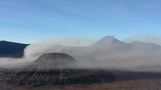 A static aerial drone shot frames Mount Bromo in the foreground as a puff of smoke from Mount Semeru drifts across the sky behind, East Java, Indonesia. Cinematic locked-off volcano b-roll.