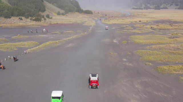 Aerial drone follows then sweeps in front of two 4x4 jeeps crossing the grey volcanic sand at Mount Bromo, ending on more jeeps parked among horses, mountains behind, East Java, Indonesia.