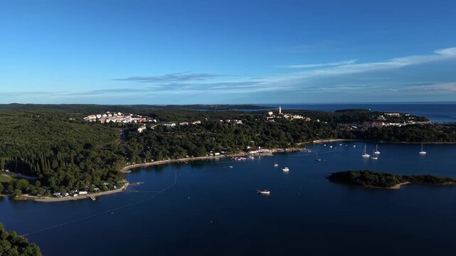 Vrsar resort town nestles among pine trees on hillside, hilltop church tower overlooking sailboats anchored in calm blue bay dotted with small wooded islets. Croatia. Aerial drone approaching shot