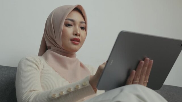 Beautiful Asian Muslim woman wearing hijab is using tablet device while sitting on a sofa.