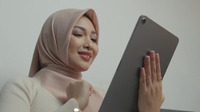 Cheerful Asian Muslim woman wearing hijab is using tablet device while sitting on a sofa.