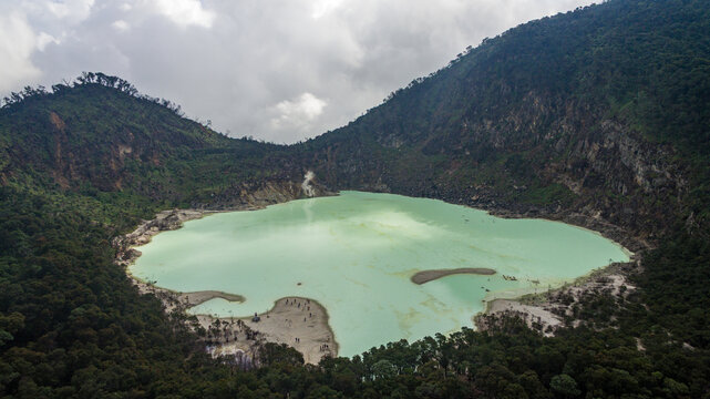 Aerial view of the White Crater volcanic lake with its turquoise water surrounded by lush forest-covered mountains and rocky slopes in Bandung, West Java, Indonesia.