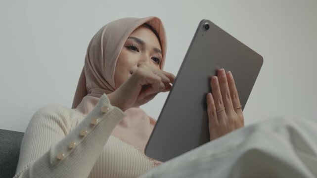 Pretty young Asian Muslim woman wearing hijab is using tablet device while sitting on a sofa.