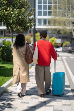 Fashionable married couple african american man and woman travelers walking on city street with suitcase, stylish black male and female partners walk together in downtown with luggage on way to hotel.