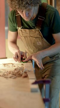 Carpenter processes future wooden legs of chair, shaping product and checks evenness of surface, small timber furniture production workshop, male woodworker using hand plane tool to model new item.