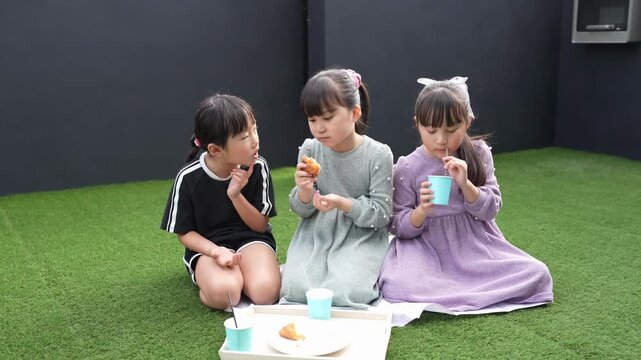Three children eat snacks outdoors on green grass. They sit close, focused on their treats and cups. Their expressions show quiet enjoyment and contentment