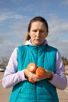 Woman holding orange dumbbells in both hands during outdoor sport workout session on sunny day in coastal parking lot. Sport exercise portrait shows focus and concentration on fitness activity.