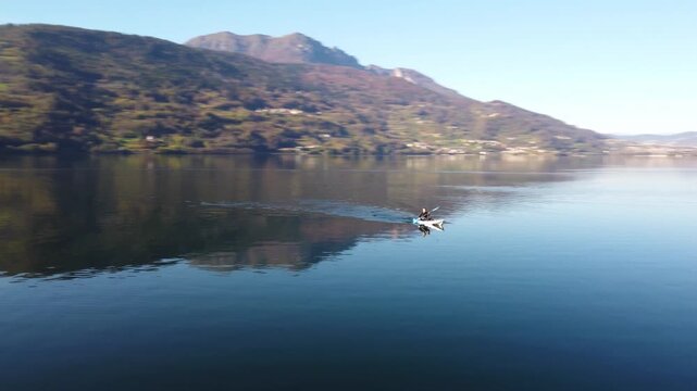Aerial 4K drone video of a person canoeing on the calm, reflective waters of Lake Caldonazzo in Trentino, Italy, surrounded by beautiful Alpine mountains.