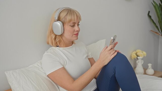Woman tapping smartphone screen with fingers, wearing silver overear headphones and white tee while seated on bed in a studio set; relaxation calm.