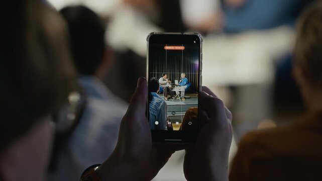 Audience member holding smartphone, recording live interview with two speakers on stage during public event. Video is perfect for mobile journalism, event marketing, or content creation ads