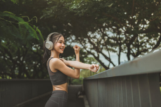 Happy Asian woman wearing headphones and stretching arms during outdoor workout in green park, smiling female athlete listening to music and warming up before running for fitness, healthy wellness.