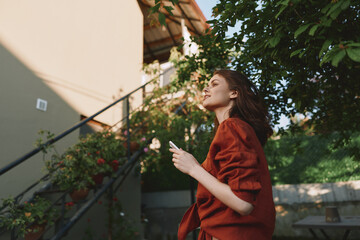 Fototapeta premium A woman in a red coat stands outdoors, looking up with a phone in hand. A calm, sunny day frames a casual street style and reflective mood