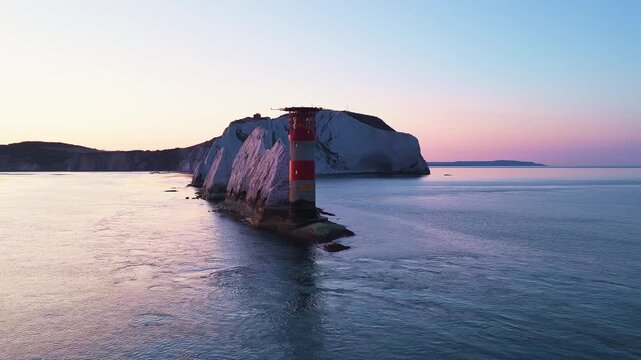 4K drone footage flying past The Needles Lighthouse at sunrise, revealing the iconic chalk stacks and western Isle of Wight headland beneath golden morning skies.