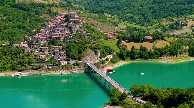 Castel di Tora Turano lake 70mm drone shot with bridge center alignment. Compressed aerial view of medieval hilltop village houses cascading down to emerald water no sky b-roll establishing