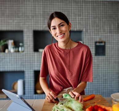 Happy young latina woman having fun preparing food and looking for recipes online using a tabletor laptop in kitchen