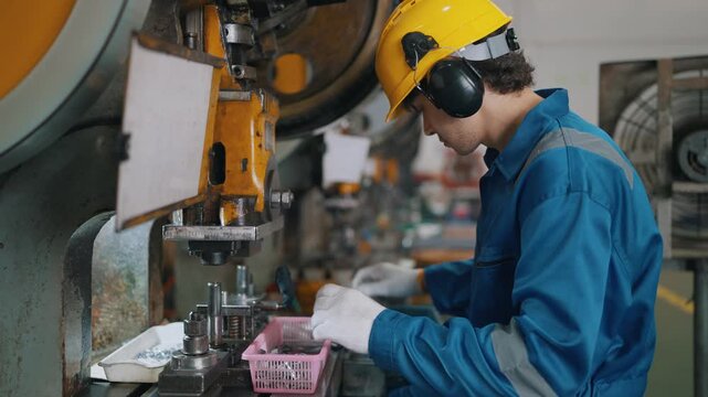 Worker wears hard hat and ear protection. Hands operate machine with precision and focus. Industrial setting shows metalworking in a factory. Scene reflects diligence and safety in manufacturing