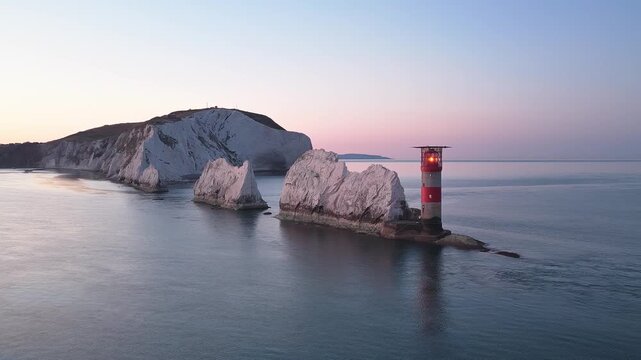 Slow panning drone shot revealing a golden dawn sky over the Needles lighthouse, chalk rocks, and the coastal cliffs of the Isle of Wight on a calm morning. 4K aerial.