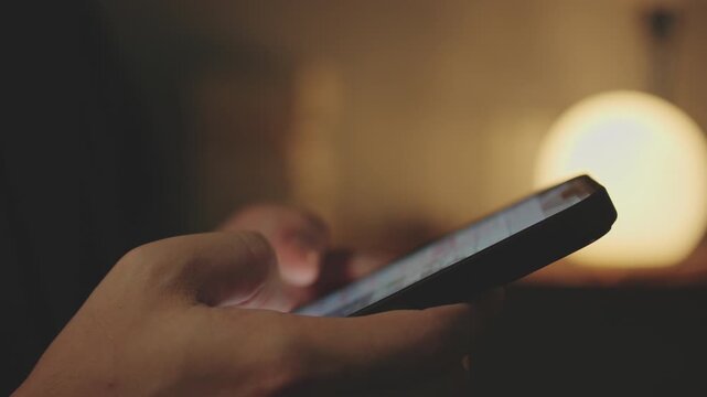 Close-up of Hand Holding and Scrolling Smartphone Screen in Dim Warm Light Room at Night.