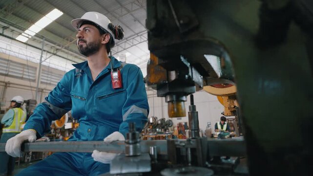 Worker in factory wears hard hat and gloves. He handles metal parts with focused, concentrated expression. Large machine dominates foreground with industrial machinery behind