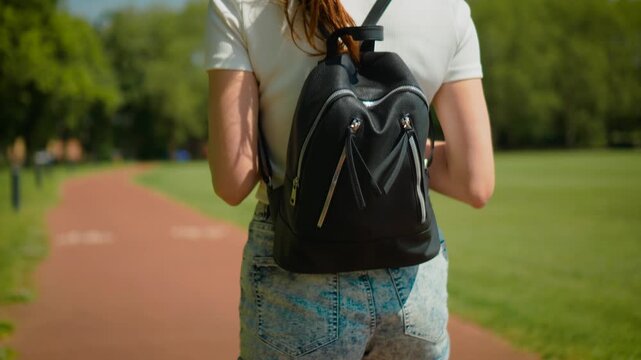 Female student carrying leather backpack while walking through athletics stadium outdoors. Healthy lifestyle and everyday movement on jogging track surrounded by green trees and open space. Urban