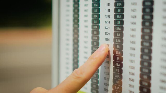 Checking bus timetable at public transport stop on empty urban street during daytime. Finger following schedule board with route numbers and departure times for city commuting. Public transportation