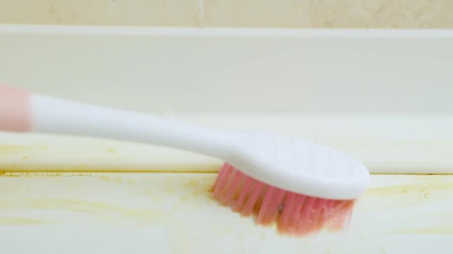 Gloved hand scrubbing bathtub seam with toothbrush and suds vigorous motion blurring bristles foam mixing with yellow residue protective glove suggests professional cleaning action