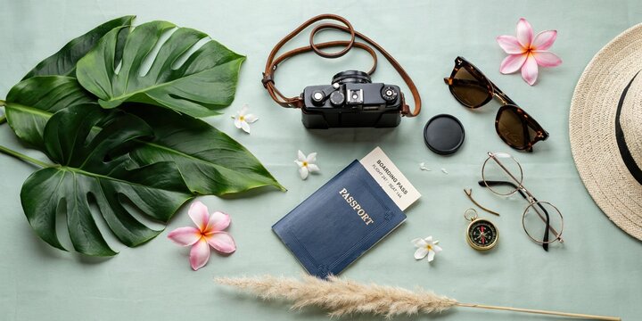 Flat lay of travel essentials with passport and vintage camera on green background. Tropical vacation concept with monstera leaves and frangipani flowers