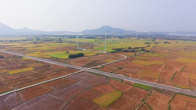 At Dam Nai Wind Farm in Ninh Thuan, wind turbines turn slowly above large fields. The landscape shows rows of planted crops and mountains in the background. A clear sky adds to the view.