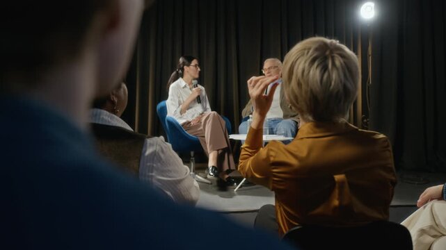 Adult Caucasian woman raising hand in audience, standing up to ask question during panel discussion with speakers on stage. Ideal for corporate training or educational conference videos