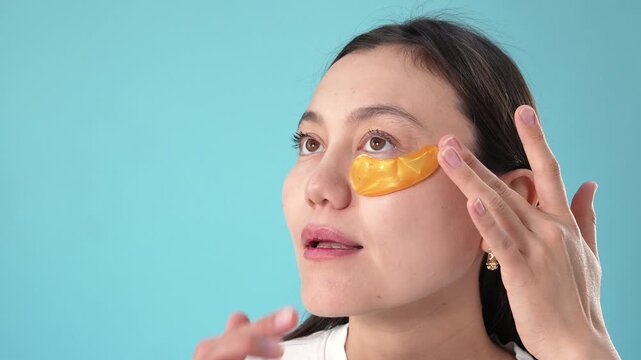 woman sequentially applying eye patches against a solid light blue background, calm and focused frontal view with slight upward angle, beauty care and skincare.