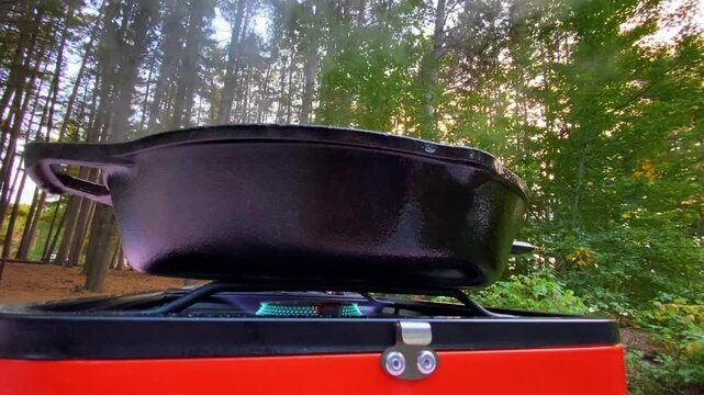 Close-up of a cast iron pan steaming over a propane flame at an outdoor cooking setup, with trees and nature visible in the background.