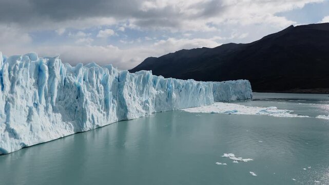 Perito Moreno Glacier stretches across Lago Argentino in Los Glaciares National Park, Santa Cruz, Argentina, as towering blue ice cliffs dominate the scene, drone lateral slide approaching ice wall
