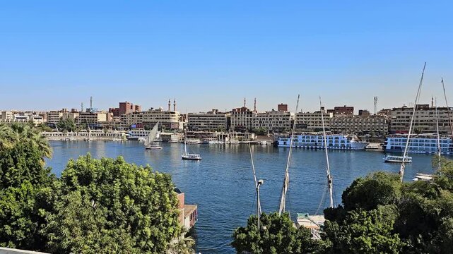 Scenic Nile River view from Elephantine Island at Aswan, Egypt, with tourist ferry boats, granite rocks, lush green island, sandy desert hills and city under blue sky.