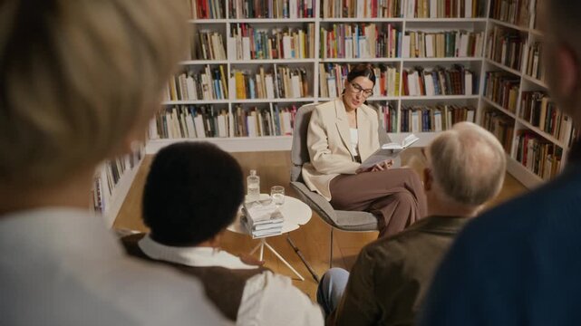 Middle aged Caucasian female author presenting book to diverse audience in library, listeners applauding speaker. Video is suitable for educational promotions or literary marketing campaigns