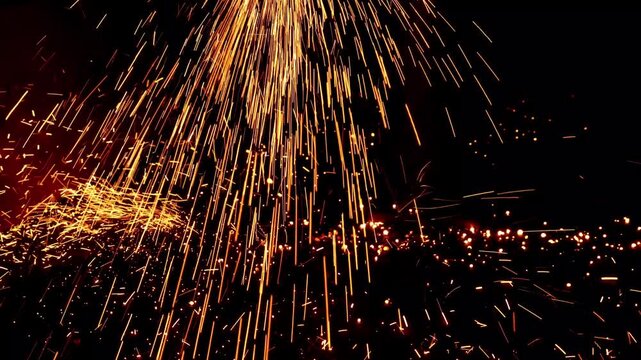Bright yellow and orange fiery sparks showering down and scattering across a dark surface during an industrial metalworking process.