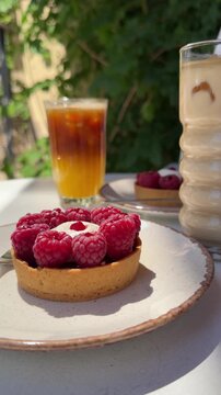 Raspberry tart with cream and fresh raspberries served on a ceramic plate in a sunny caf&eacute; garden, accompanied by iced coffee and latte on a summer outdoor table.